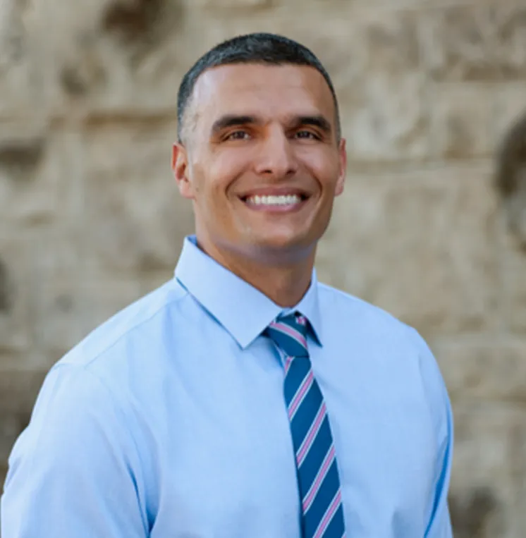 Smiling cosmetic and family dentist headshot in blue shirt and striped tie outdoors.